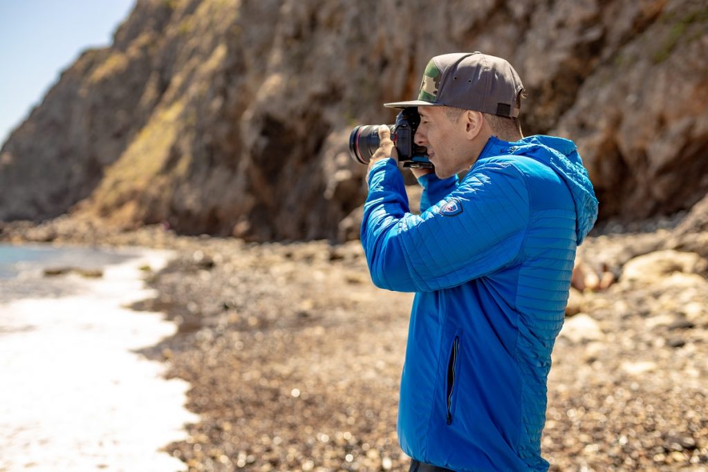man in blue K&Uuml;HL jacket holding camera on coastline