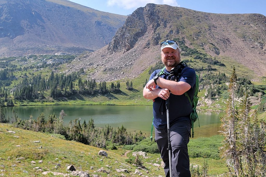 man in K&Uuml;HL shirt standing in front of the lake during daytime